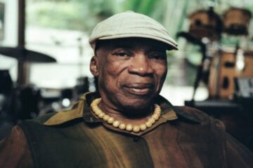 : Milton Nascimento sentado, usando camisa em tons terrosos, colar de contas e boné claro, com expressão serena em estúdio musical.