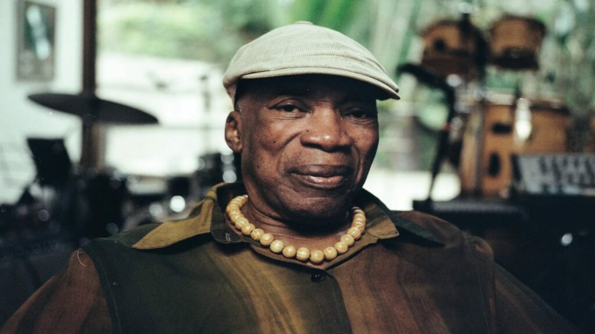 : Milton Nascimento sentado, usando camisa em tons terrosos, colar de contas e boné claro, com expressão serena em estúdio musical.