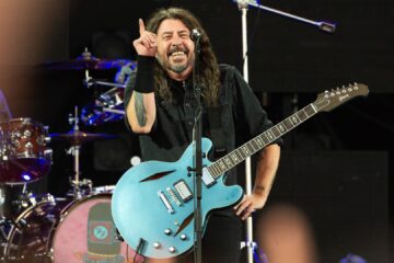 Dave Grohl sorrindo e apontando para cima enquanto toca guitarra azul em show do Foo Fighters.