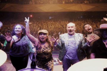 Banda Men At Work sorrindo no palco durante show lotado no Brasil.