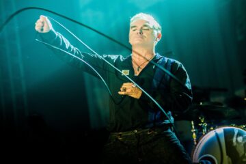 Morrissey se apresentando ao vivo com expressão intensa, olhos fechados e segurando o cabo do microfone, em show iluminado por luz azul.