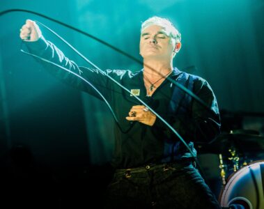 Morrissey se apresentando ao vivo com expressão intensa, olhos fechados e segurando o cabo do microfone, em show iluminado por luz azul.