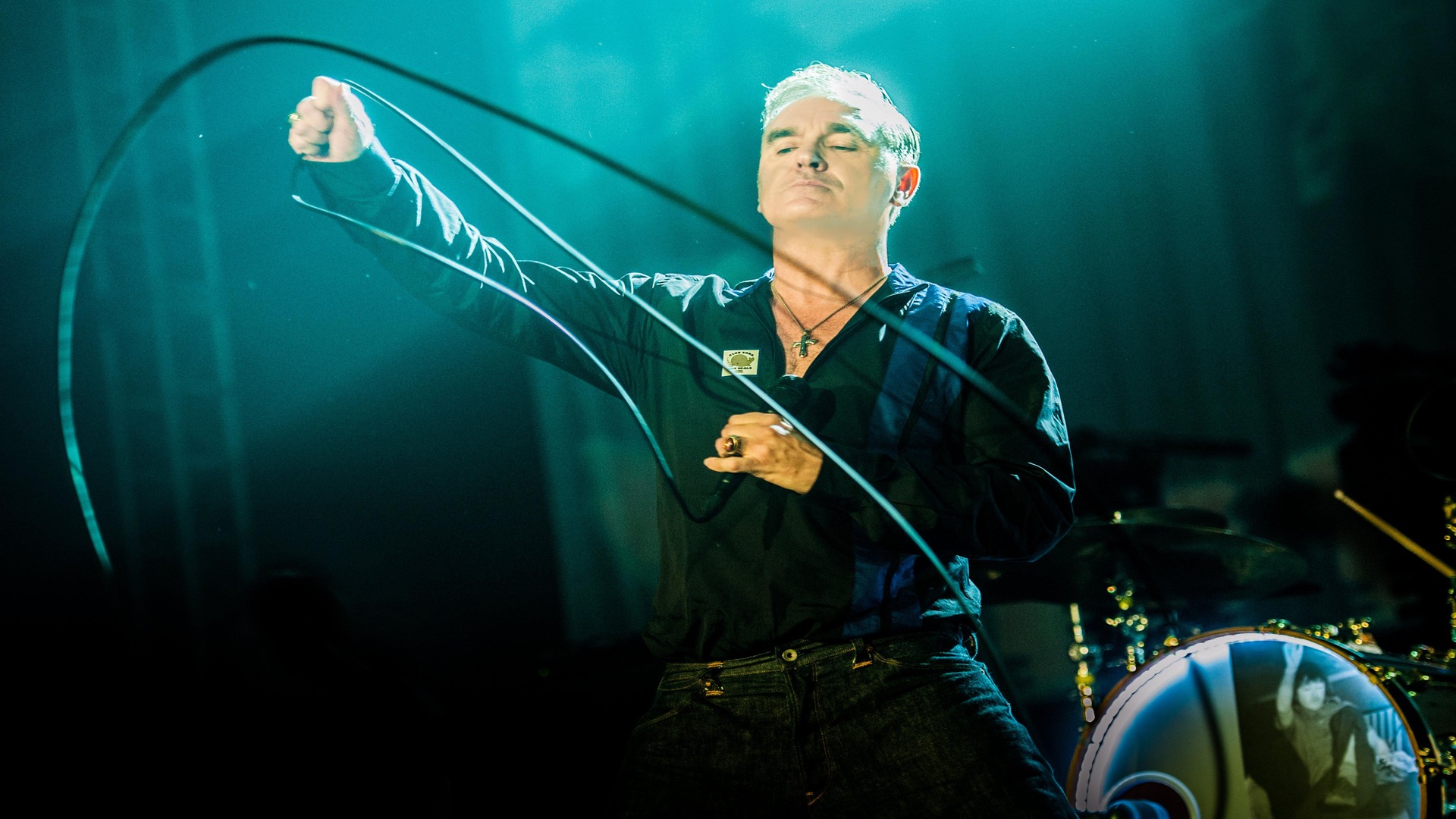 Morrissey se apresentando ao vivo com expressão intensa, olhos fechados e segurando o cabo do microfone, em show iluminado por luz azul.