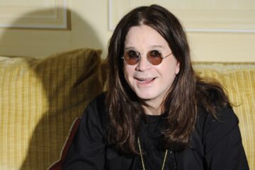 Ozzy Osbourne sorri sentado em um sofá, usando óculos redondos e roupas pretas, em retrato promocional.