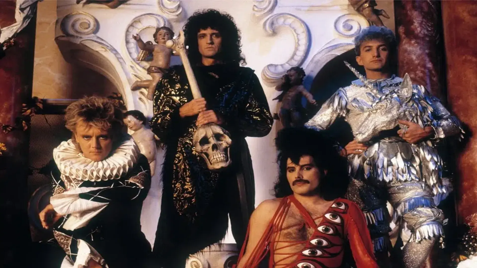 Queen em registro promocional nos anos 80, com Freddie Mercury, Brian May, Roger Taylor e John Deacon usando figurinos teatrais e posando em cenário barroco. Foto: Queen Productions Ltd.