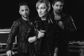 Foto em preto e branco do The Calling: Alex Band ao centro com jaqueta de couro, acompanhado por dois integrantes da banda, posando em estúdio com cases e guitarra ao fundo.