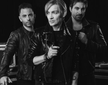 Foto em preto e branco do The Calling: Alex Band ao centro com jaqueta de couro, acompanhado por dois integrantes da banda, posando em estúdio com cases e guitarra ao fundo.