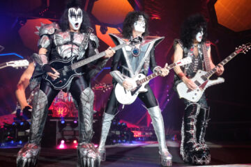 Integrantes do Kiss tocando ao vivo com seus figurinos clássicos e maquiagens icônicas durante apresentação em grande palco iluminado, com efeitos de luzes e cenário futurista ao fundo.