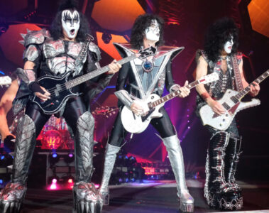 Integrantes do Kiss tocando ao vivo com seus figurinos clássicos e maquiagens icônicas durante apresentação em grande palco iluminado, com efeitos de luzes e cenário futurista ao fundo.