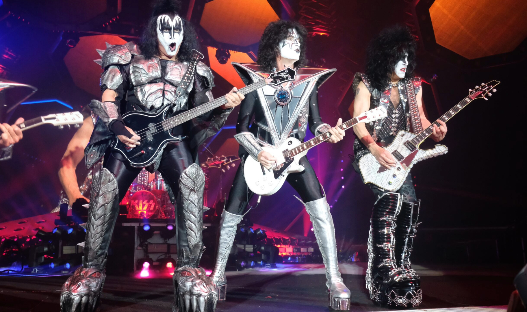 Integrantes do Kiss tocando ao vivo com seus figurinos clássicos e maquiagens icônicas durante apresentação em grande palco iluminado, com efeitos de luzes e cenário futurista ao fundo.