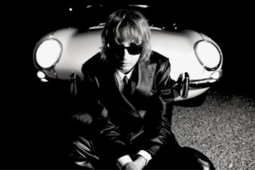 Thomas Raggi, do Måneskin, sentado em frente a um carro clássico à noite, usando terno preto e óculos escuros, em foto promocional do álbum solo Masquerade.