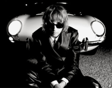 Thomas Raggi, do Måneskin, sentado em frente a um carro clássico à noite, usando terno preto e óculos escuros, em foto promocional do álbum solo Masquerade.