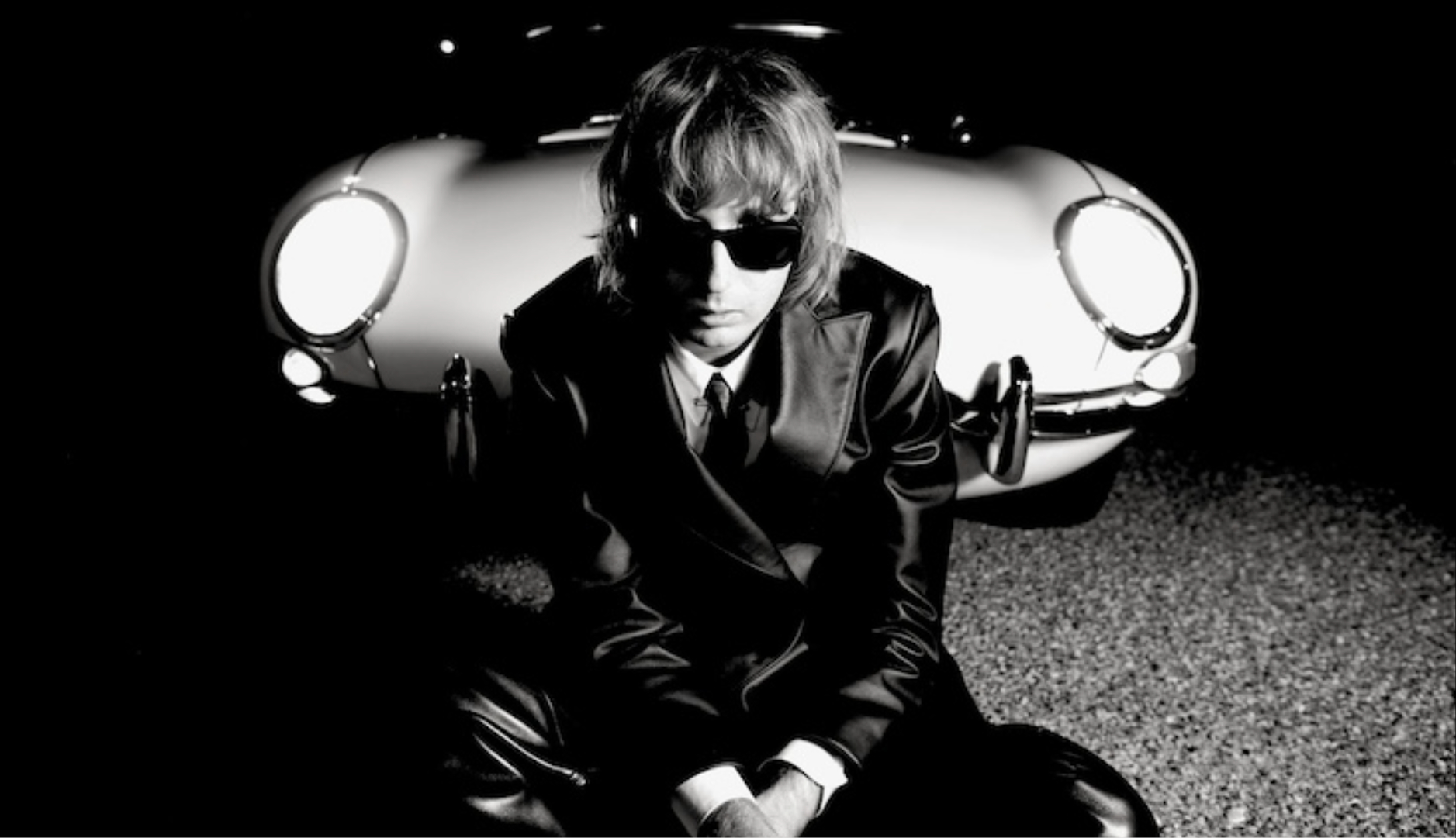 Thomas Raggi, do Måneskin, sentado em frente a um carro clássico à noite, usando terno preto e óculos escuros, em foto promocional do álbum solo Masquerade.