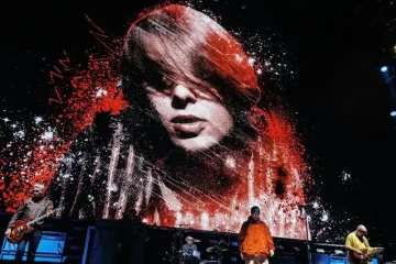 Oasis no palco em São Paulo com imagem de Mani Mounfield em vermelho e branco exibida no telão durante homenagem.