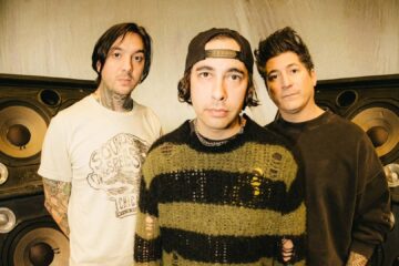 Foto promocional do Pierce The Veil: três integrantes posam em estúdio, na frente de caixas de som enormes, com visual alternativo e estilo característico da banda.