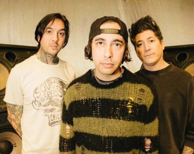 Foto promocional do Pierce The Veil: três integrantes posam em estúdio, na frente de caixas de som enormes, com visual alternativo e estilo característico da banda.