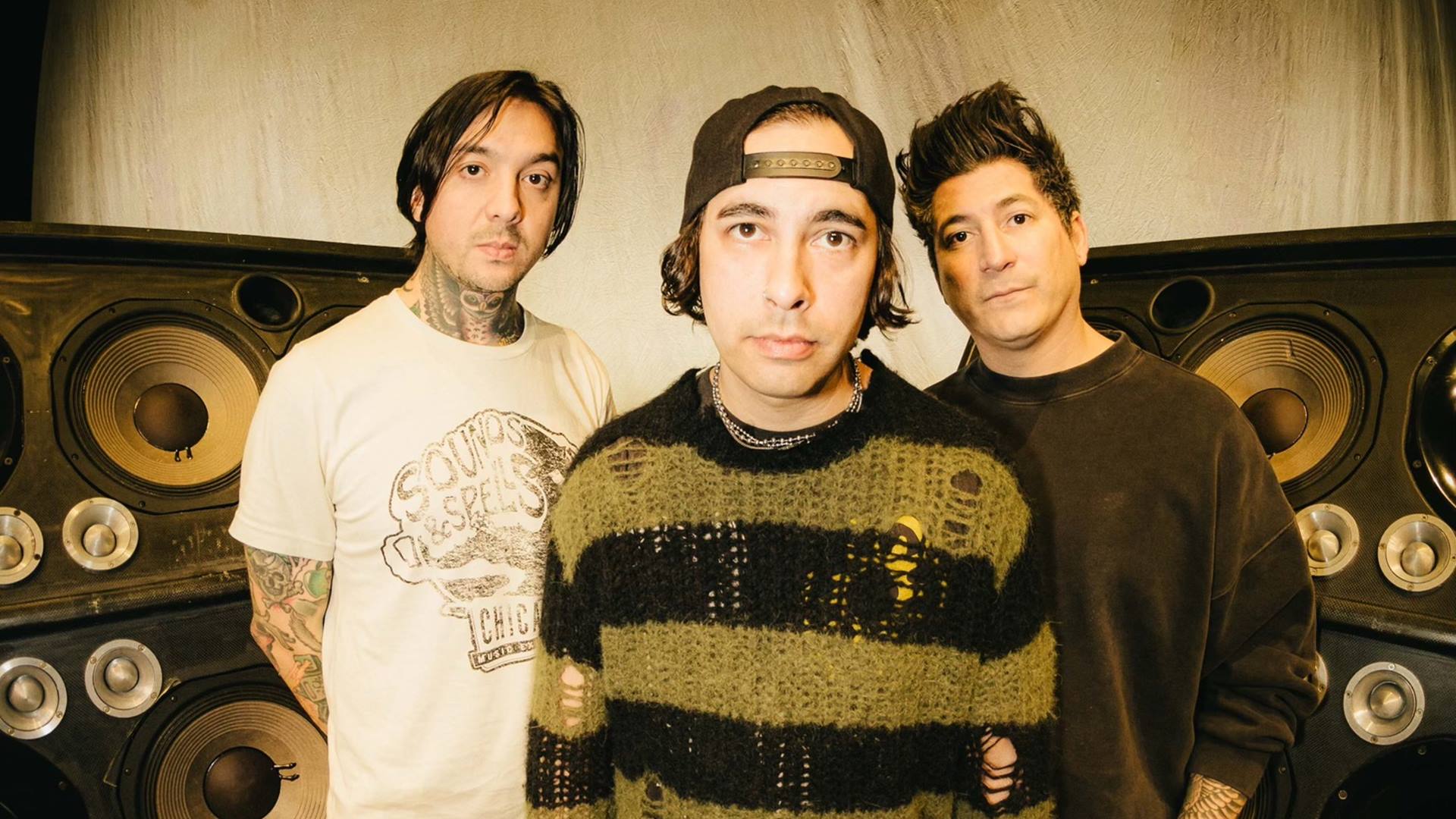 Foto promocional do Pierce The Veil: três integrantes posam em estúdio, na frente de caixas de som enormes, com visual alternativo e estilo característico da banda.