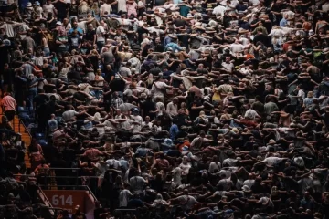 Multidão em um estádio de shows realizando o gesto Poznan: milhares de pessoas de costas para o palco, unidas pelos ombros e pulando em sincronia.