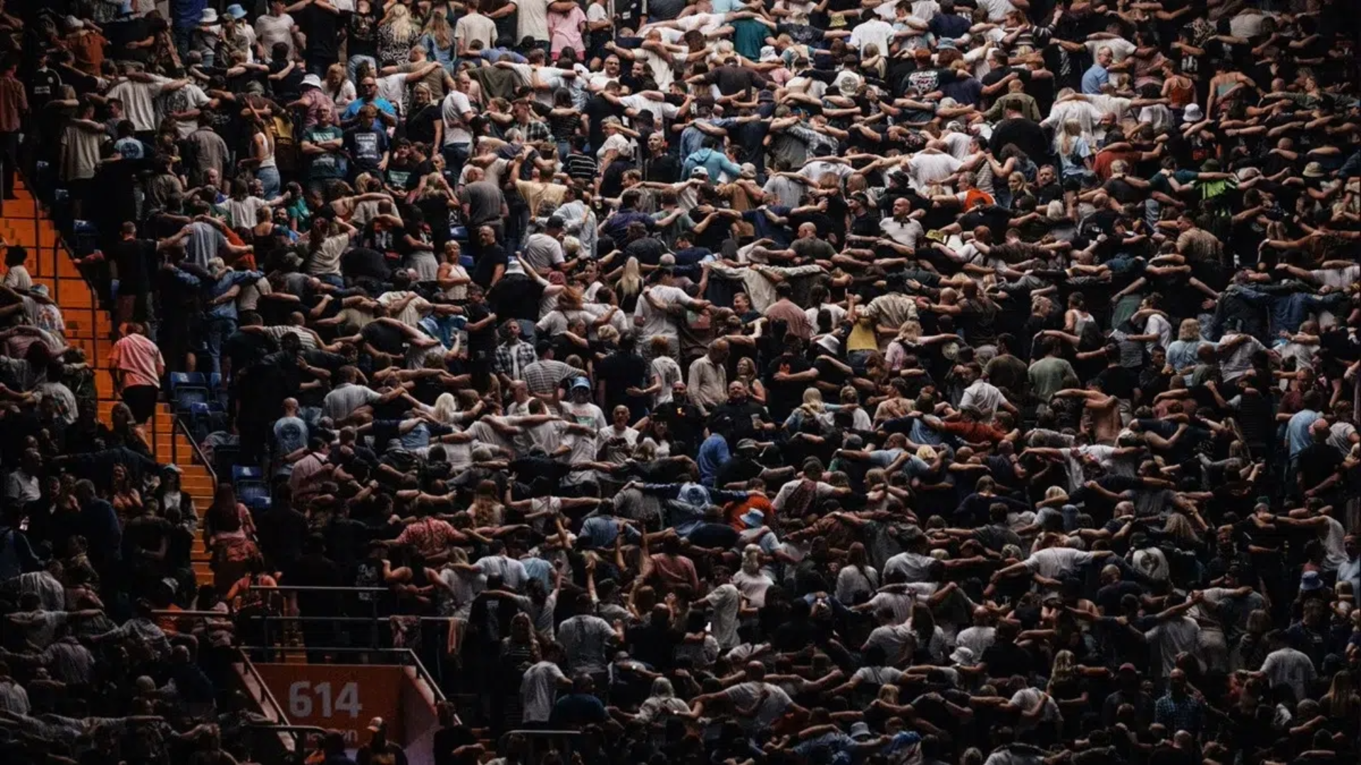 Multidão em um estádio de shows realizando o gesto Poznan: milhares de pessoas de costas para o palco, unidas pelos ombros e pulando em sincronia.