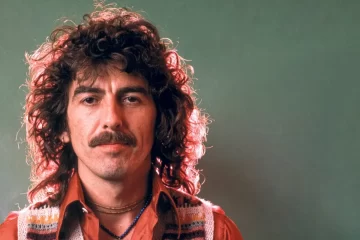 George Harrison em foto de estúdio, com cabelo encaracolado e camisa laranja, olhando diretamente para a câmera.