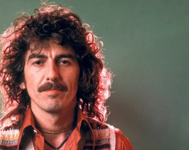 George Harrison em foto de estúdio, com cabelo encaracolado e camisa laranja, olhando diretamente para a câmera.