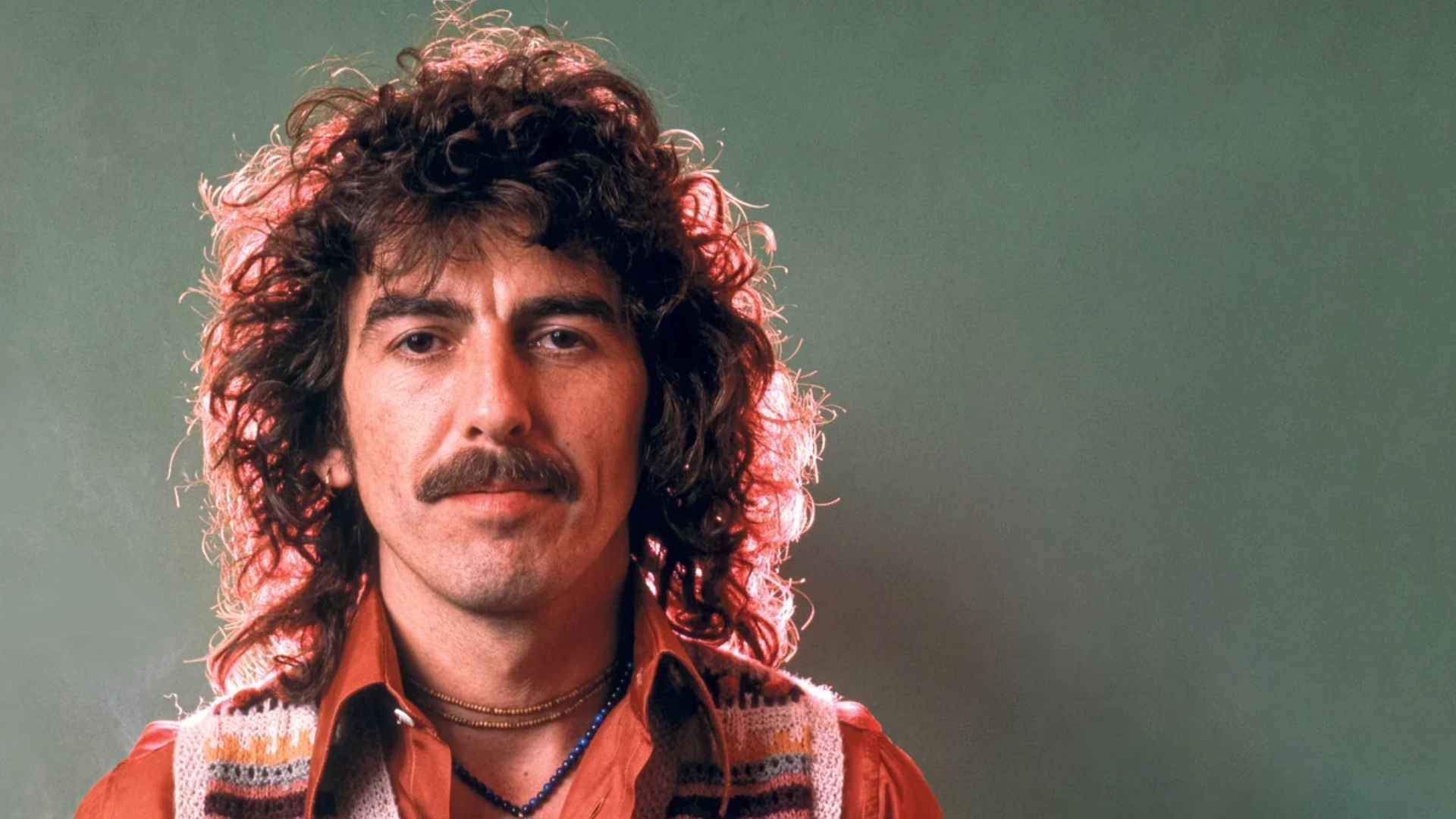 George Harrison em foto de estúdio, com cabelo encaracolado e camisa laranja, olhando diretamente para a câmera.