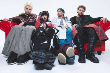 Integrantes do 5 Seconds of Summer sentados em um sofá vermelho em ensaio fotográfico promocional.