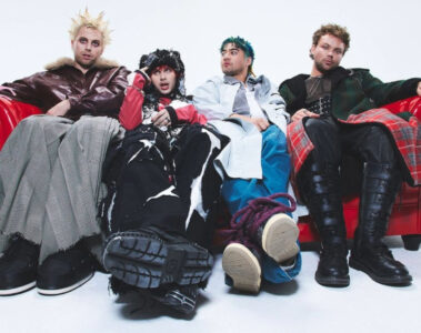 Integrantes do 5 Seconds of Summer sentados em um sofá vermelho em ensaio fotográfico promocional.