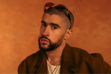 Bad Bunny posando com expressão séria em fundo laranja, usando corrente dourada e óculos sobre a cabeça.