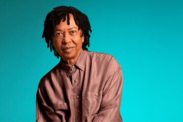 Retrato de Djavan sentado diante de um fundo azul turquesa, usando camisa e calça em tom cobre, para divulgar a turnê comemorativa de 50 anos.