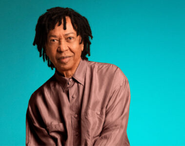 Retrato de Djavan sentado diante de um fundo azul turquesa, usando camisa e calça em tom cobre, para divulgar a turnê comemorativa de 50 anos.