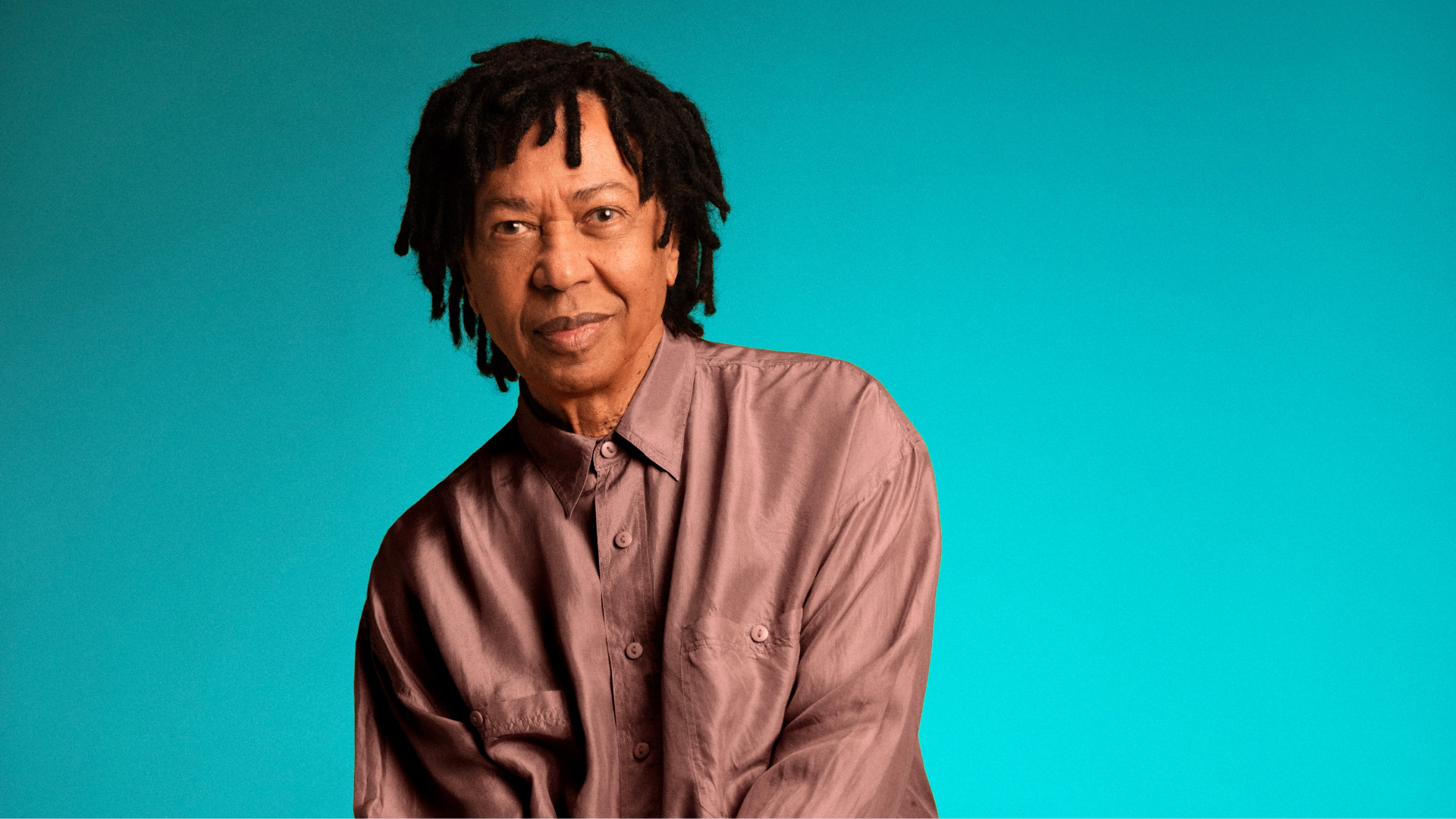 Retrato de Djavan sentado diante de um fundo azul turquesa, usando camisa e calça em tom cobre, para divulgar a turnê comemorativa de 50 anos.