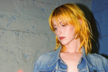 Hayley Williams com cabelo amarelo dourado olhando para baixo em um ambiente urbano.