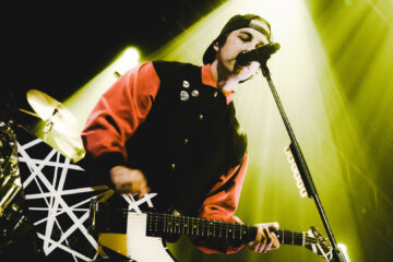 Vic Fuentes, vocalista do Pierce the Veil, canta e toca guitarra durante show em Curitiba, iluminado por luzes amarelas no palco. Foto de Ravi Isfer para a Moodgate.