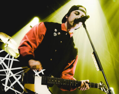 Vic Fuentes, vocalista do Pierce the Veil, canta e toca guitarra durante show em Curitiba, iluminado por luzes amarelas no palco. Foto de Ravi Isfer para a Moodgate.