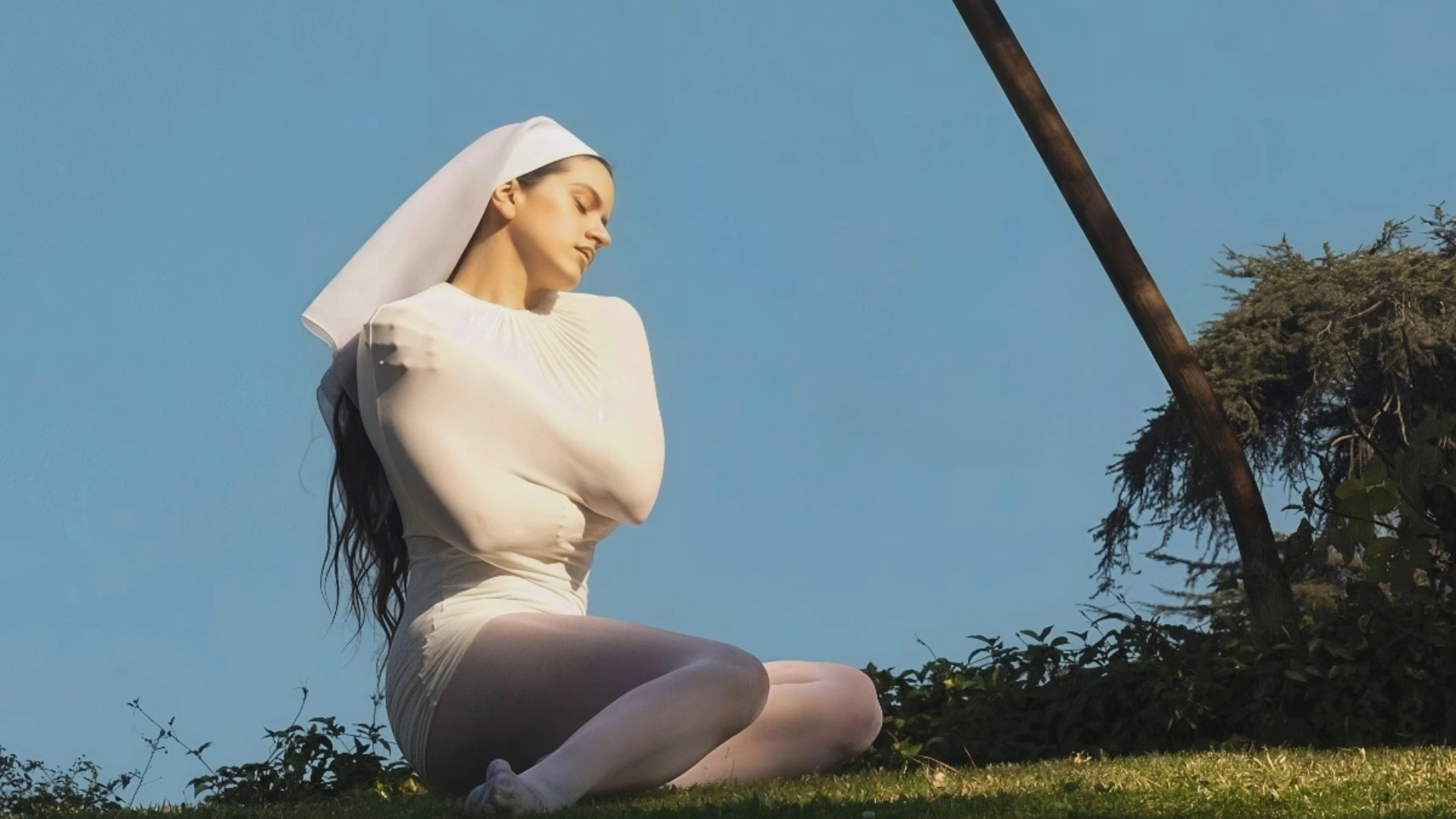 Rosalía sentada em um gramado, vestindo figurino branco, em imagem promocional do álbum “LUX”.