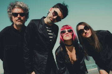 Integrantes da banda Supercombo posando em uma praia, usando roupas escuras e óculos escuros, em foto promocional do álbum Caranguejo.