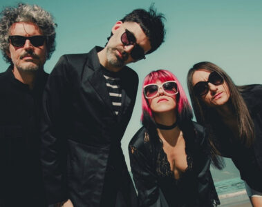 Integrantes da banda Supercombo posando em uma praia, usando roupas escuras e óculos escuros, em foto promocional do álbum Caranguejo.