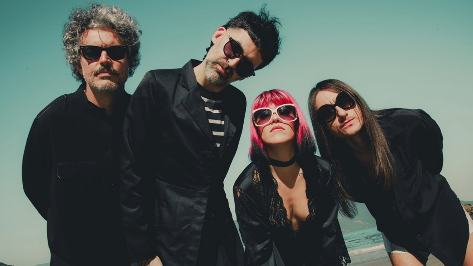 Integrantes da banda Supercombo posando em uma praia, usando roupas escuras e óculos escuros, em foto promocional do álbum Caranguejo.