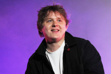 Lewis Capaldi sorrindo no palco durante apresentação ao vivo, com iluminação roxa ao fundo.