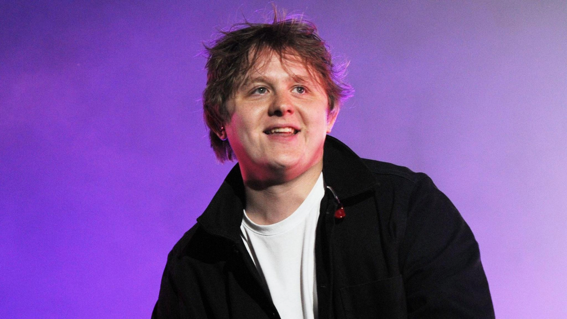 Lewis Capaldi sorrindo no palco durante apresentação ao vivo, com iluminação roxa ao fundo.