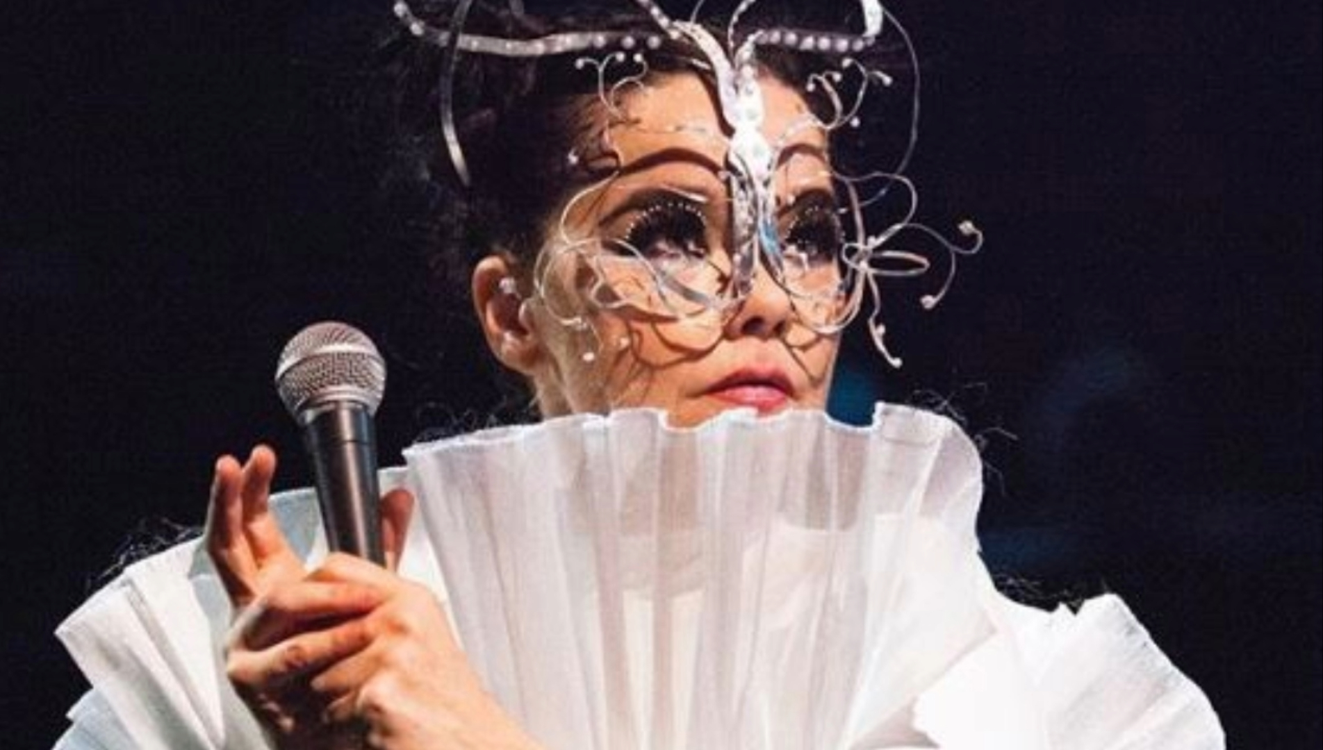 Björk segurando um microfone e usando uma máscara artística detalhada, durante apresentação ao vivo.