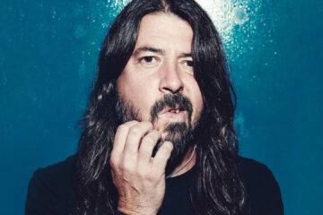 Dave Grohl em um retrato de close-up, com expressão séria e a mão tocando a barba, posando diante de um fundo azul.