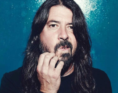 Dave Grohl em um retrato de close-up, com expressão séria e a mão tocando a barba, posando diante de um fundo azul.