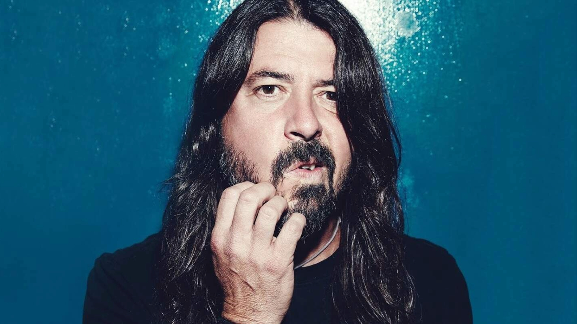 Dave Grohl em um retrato de close-up, com expressão séria e a mão tocando a barba, posando diante de um fundo azul.