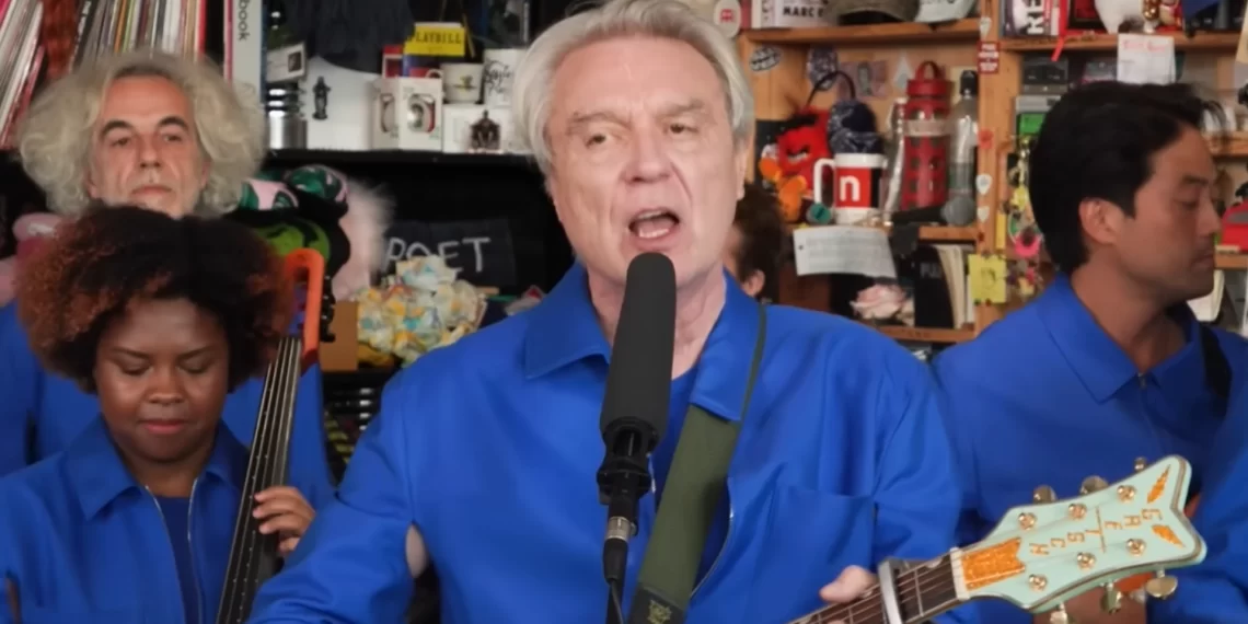 David Byrne cantando e tocando guitarra durante seu Tiny Desk Concert na NPR, acompanhado por sua banda, foto via YouTube.