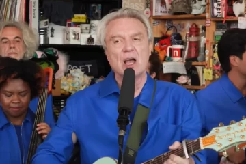 David Byrne cantando e tocando guitarra durante seu Tiny Desk Concert na NPR, acompanhado por sua banda, foto via YouTube.