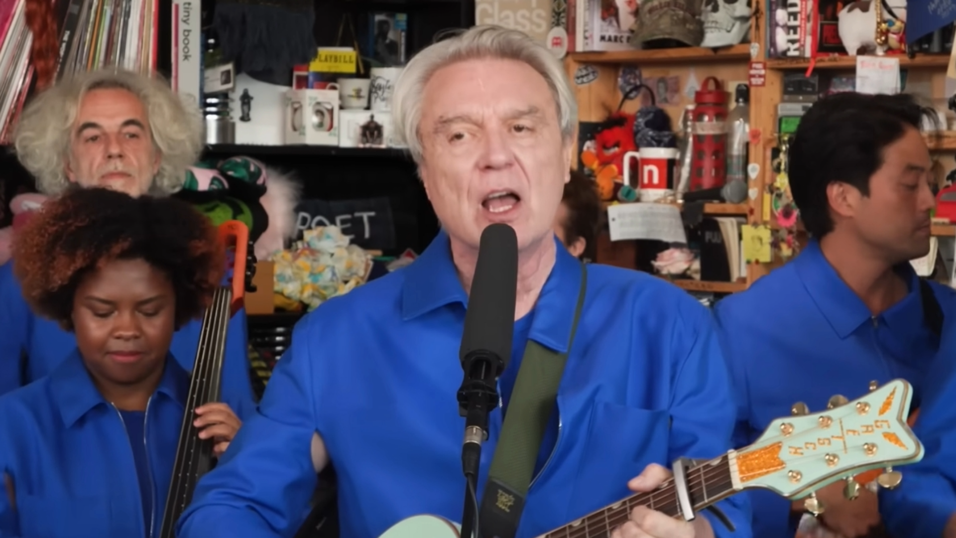 David Byrne cantando e tocando guitarra durante seu Tiny Desk Concert na NPR, acompanhado por sua banda, foto via YouTube.