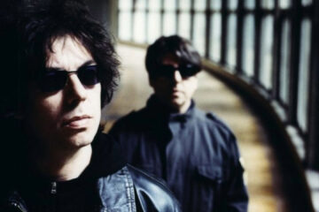 Duo do Echo & The Bunnymen usando óculos escuros em corredor iluminado por janelas, em imagem promocional.