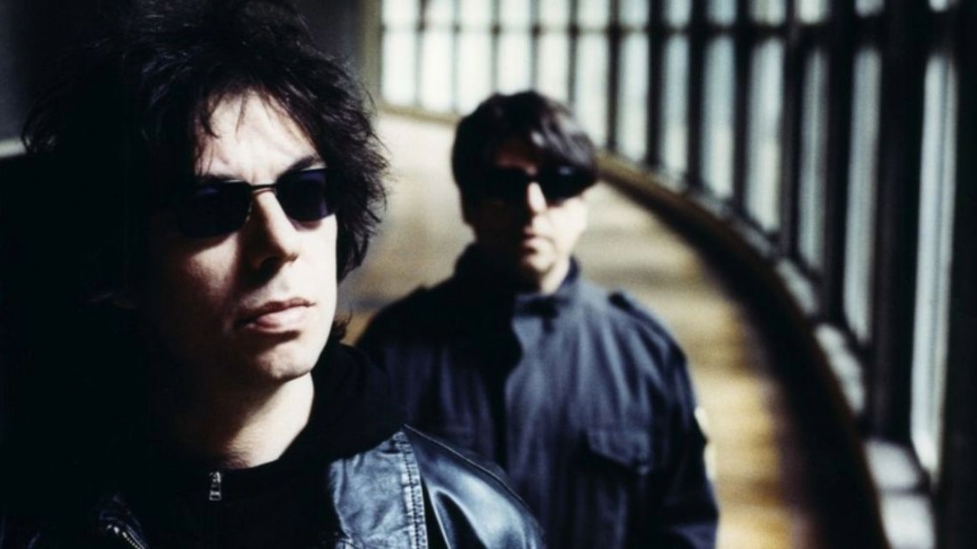 Duo do Echo & The Bunnymen usando óculos escuros em corredor iluminado por janelas, em imagem promocional.